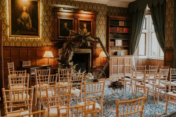 Intimate ceremony space with wooden chairs, vintage wallpaper, and a cozy fireplace, complemented by artistic décor and bookshelves.
