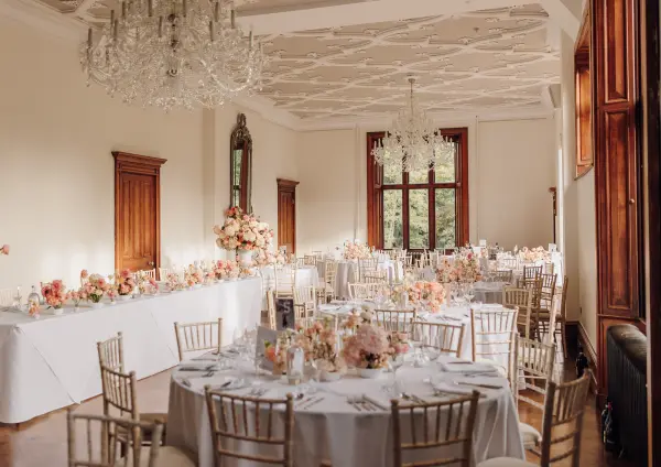 Elegant banquet room featuring round tables adorned with soft floral arrangements, crystal chandeliers, and large windows for natural light.