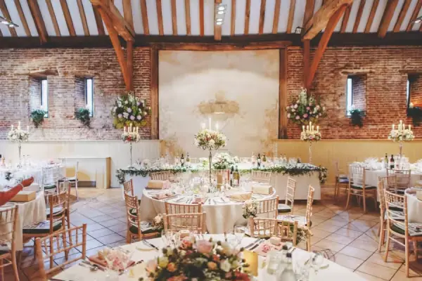 Rustic barn interior features exposed beams, elegant round tables adorned with floral centerpieces, and charming candlelit decor.