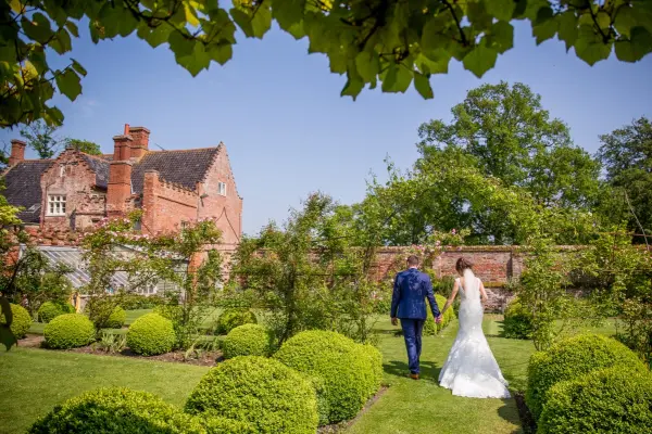 Couple walks hand in hand through lush gardens with manicured hedges and a stunning rustic estate in the background, perfect for a romantic wedding.