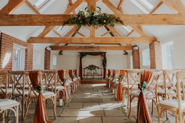 Charming ceremony space featuring rustic wooden beams, lined with cross-back chairs adorned with warm fabric sashes and floral accents.