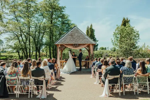Outdoor wedding ceremony under a charming gazebo, surrounded by lush greenery and seated guests, creating an intimate atmosphere.