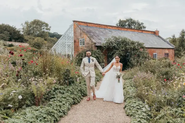 Couple walks hand in hand through lush gardens, with a charming rustic building and glass greenhouse in the background. Perfect for outdoor ceremonies.