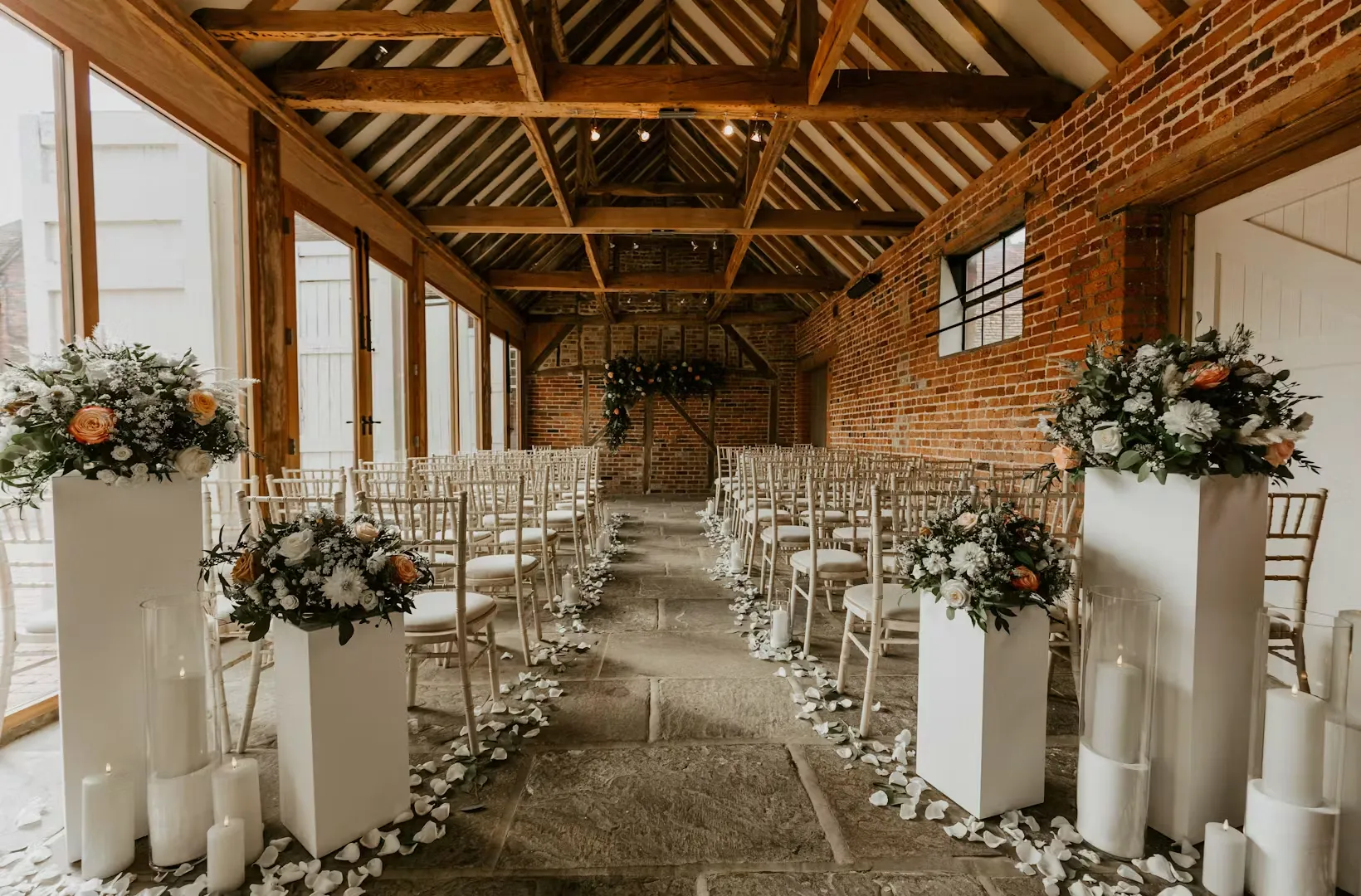 Charming indoor ceremony space featuring exposed wood beams, elegant floral arrangements, and rows of chairs on a stone floor.