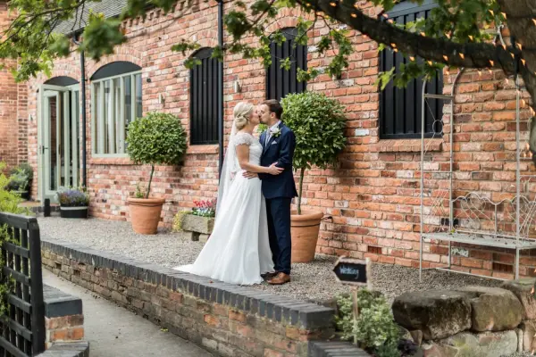 Intimate outdoor setting featuring a couple embracing near a rustic brick wall, adorned with greenery and potted plants, perfect for romantic weddings.