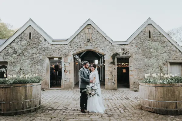 Couple embraces in front of a rustic stone building, featuring wooden planters and charming archways for an enchanting wedding backdrop.