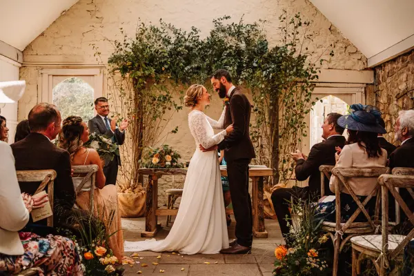 Intimate indoor ceremony space with stone walls, lush greenery, and wooden chairs, featuring a joyful couple exchanging vows.