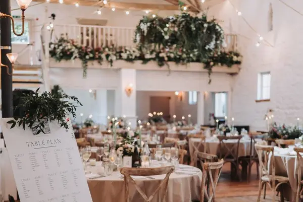 Charming rustic barn interior featuring elegantly set round tables, lush greenery, and soft string lights for a romantic atmosphere.