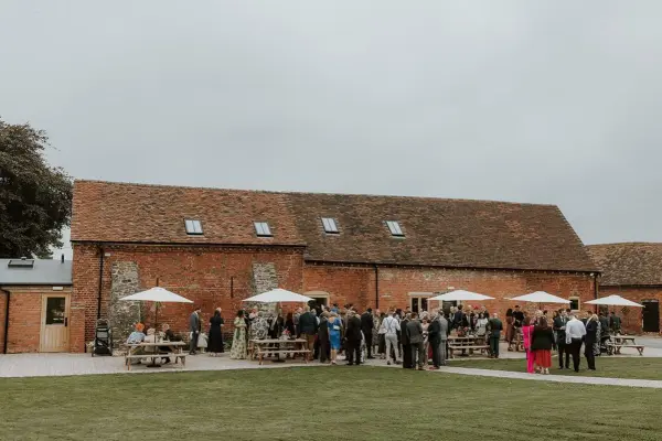 Rustic barn wedding venue featuring a spacious outdoor lawn, surrounded by guests enjoying drinks under shaded umbrellas on a cloudy day.