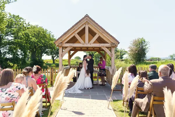 Couple exchange vows under a charming wooden gazebo, surrounded by lush greenery and seated guests, creating a romantic outdoor ceremony atmosphere.