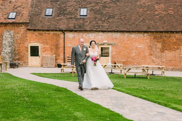 Bride and her father walk down a stone path toward a rustic barn, surrounded by green grass and wooden seating areas.