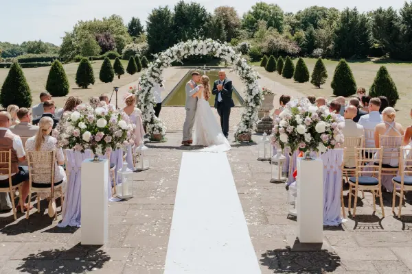Intimate outdoor ceremony with a floral arch, white aisle, and elegant arrangements, surrounded by greenery and guests in a scenic setting.