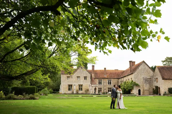 Couple embraces on a lush green lawn in front of a historic stone manor, framed by leafy trees, perfect for outdoor ceremonies.