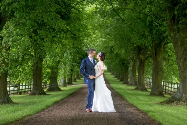 A couple embraces on a tree-lined pathway, featuring lush greenery and a charming dirt road, perfect for romantic wedding photos.