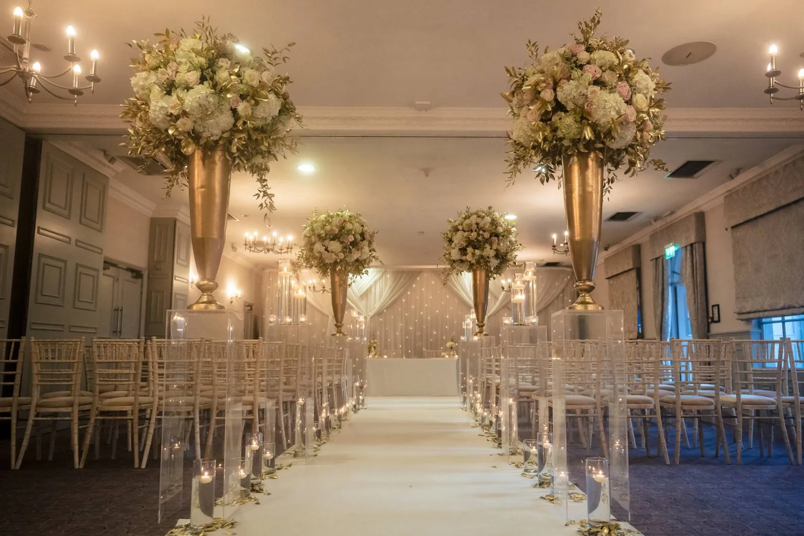 Elegant indoor wedding ceremony space featuring tall floral arrangements, golden vases, and a candlelit aisle leading to the altar.