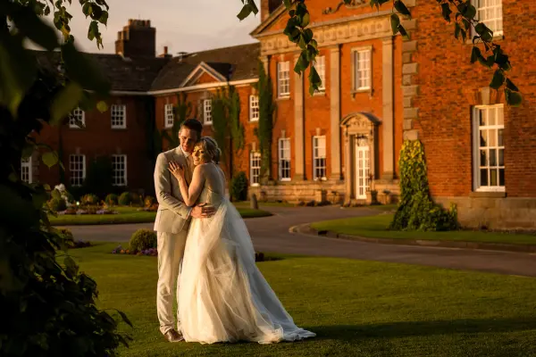 Couple embraces on manicured lawn, with a stunning red-brick mansion in the background, perfect for elegant wedding celebrations.