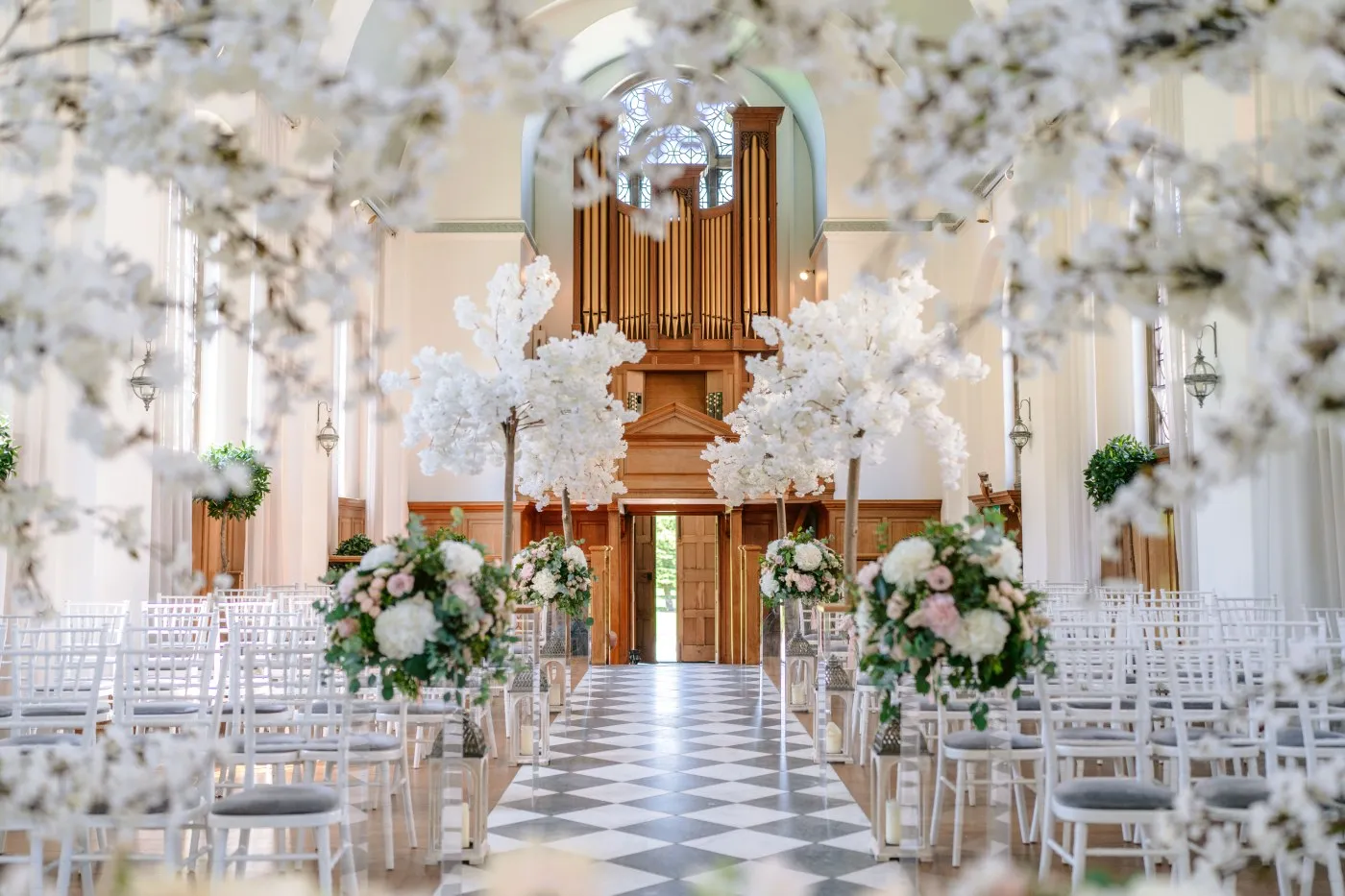 A stunning ceremony space with elegant white floral arrangements, a checkered floor, and a grand wooden altar in a light-filled setting.