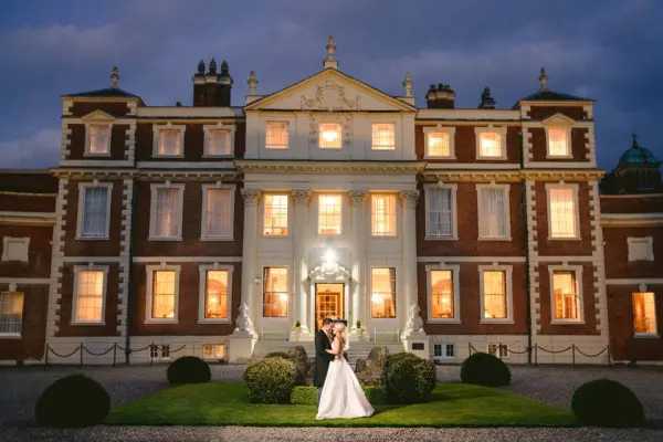 A couple stands affectionately in front of a grand manor, beautifully illuminated, showcasing elegant architecture and manicured gardens.