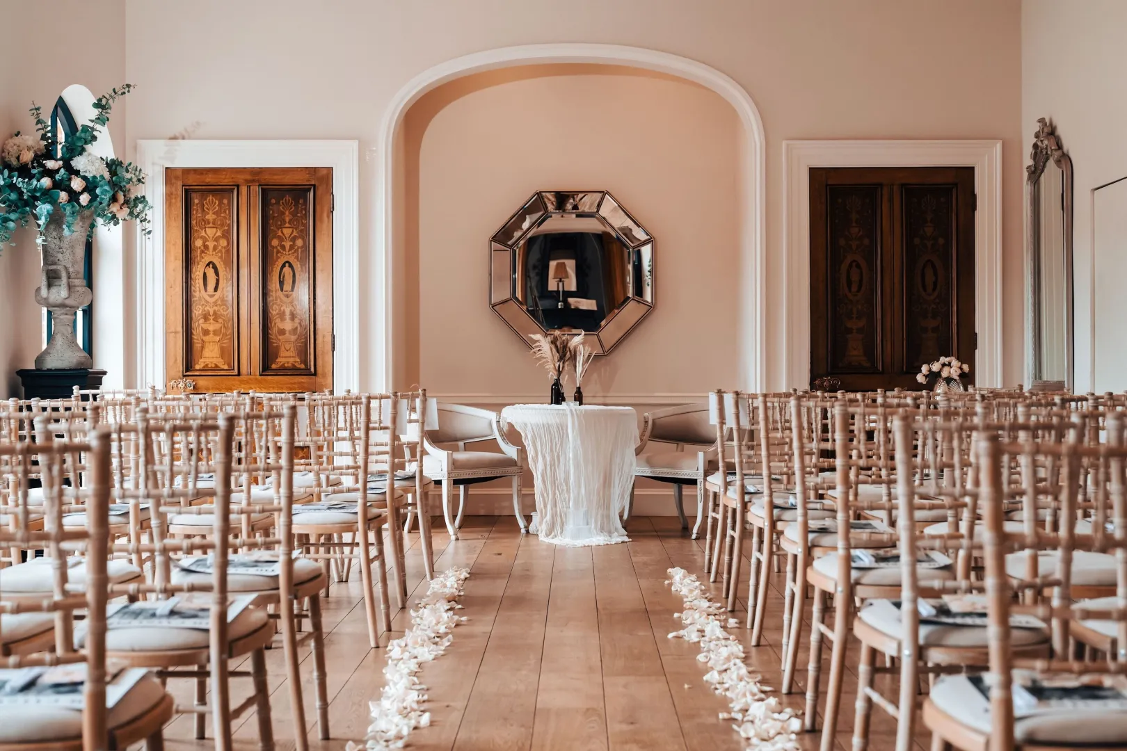 Elegant ceremony space featuring wooden floors, chiavari chairs, and a stylish backdrop with an ornate mirror and floral accents.