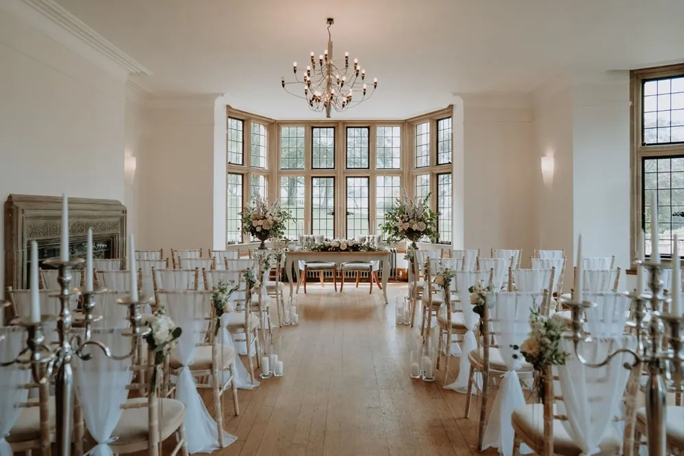 Elegant ceremony space featuring white chairs, floral arrangements, a dramatic chandelier, and large windows providing natural light.