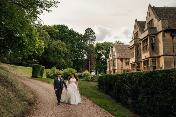 A couple strolls along a gravel path, surrounded by lush greenery and a charming historic venue with elegant architecture.