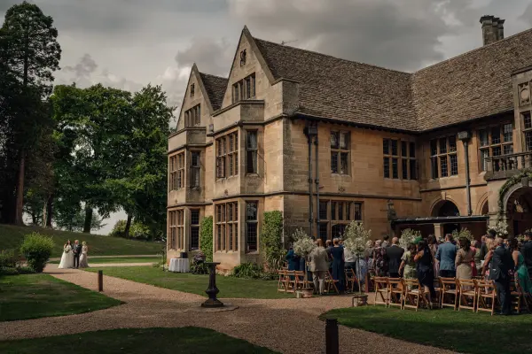 Charming outdoor wedding ceremony set in front of a grand stone manor, surrounded by lush greenery and attentive guests.