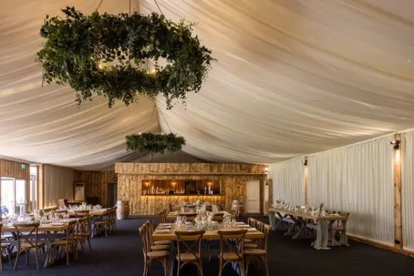 Spacious indoor reception area with elegant draped ceiling, rustic wood bar, and charming wooden tables set for a wedding celebration.