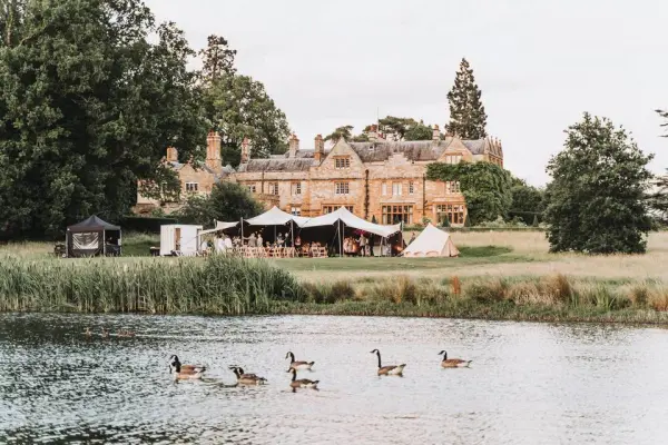 Charming lakeside venue featuring a historic manor, elegant marquees, and rustic beauty, surrounded by lush greenery and grazing geese.