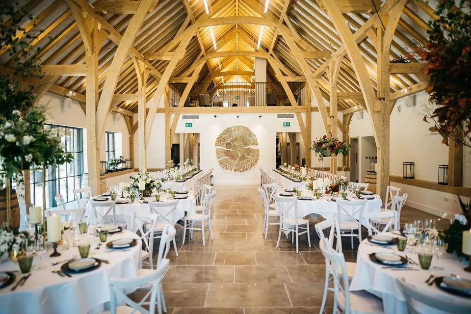 Bright, spacious barn venue featuring wooden beams, elegant tables set for a wedding, and lush greenery accents throughout.