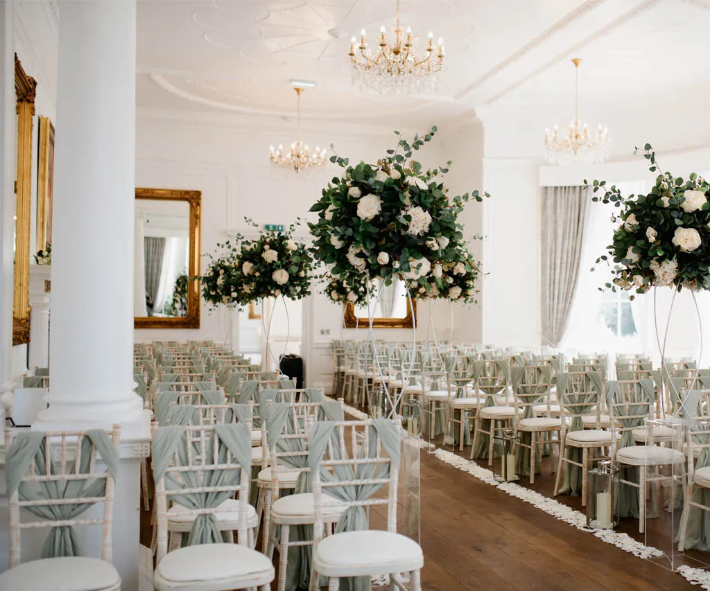 Elegant indoor ceremony space featuring white chairs adorned with soft fabrics, lush floral arrangements, and grand chandeliers.