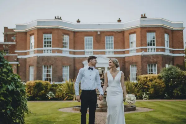 A bride and groom stroll hand in hand through manicured gardens, with a charming historic brick mansion in the background.