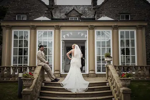 A bride in a flowing gown poses by a grand entrance with large windows, while a groom sits on stone steps, surrounded by greenery.