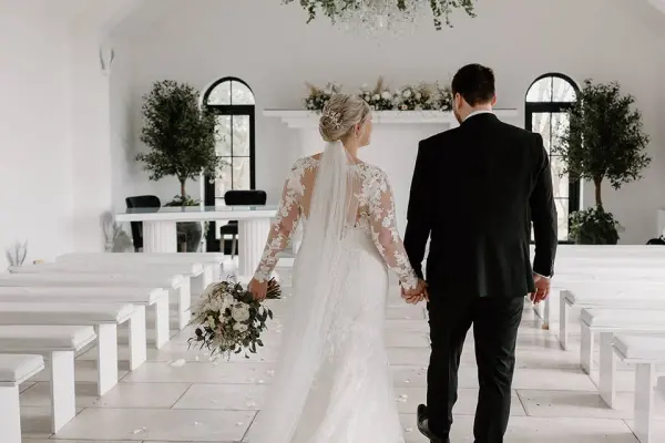 Couple walks hand in hand down a bright, modern aisle surrounded by white benches and lush greenery in an elegant ceremony space.