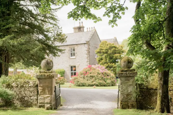 Lush greenery frames a charming stone house, with elegant gate posts leading to a picturesque garden, perfect for romantic outdoor weddings.