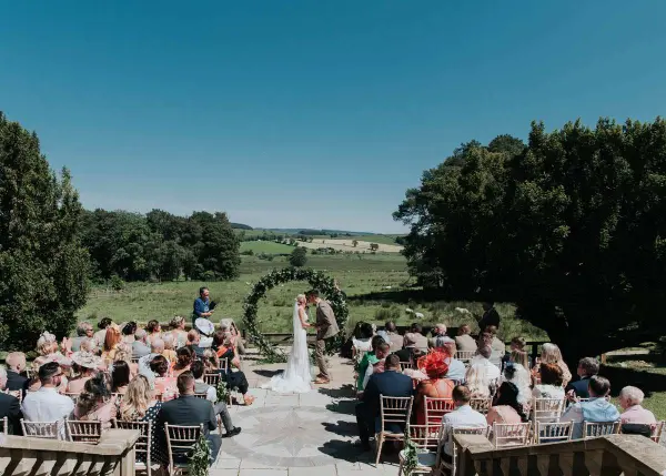 Sunlit outdoor ceremony with guests seated on stone steps, surrounded by lush greenery and scenic countryside views. Elegant floral arch enhances the setting.