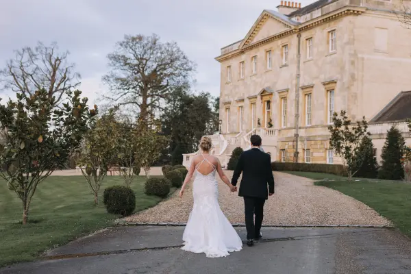 Couple walks hand in hand down a scenic path, leading to an elegant historic mansion surrounded by lush greenery and manicured gardens.