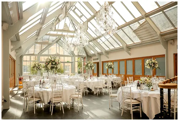 Bright and elegant glass-walled venue featuring decorated round tables, sparkling chandeliers, and lush floral arrangements. Ideal for a romantic reception.