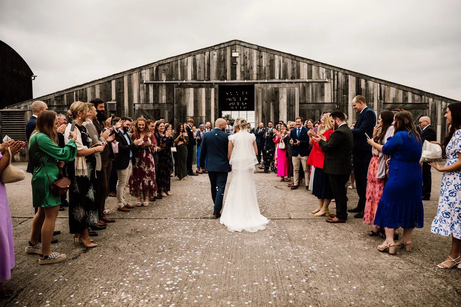 Couple walks hand-in-hand down a gravel path, flanked by guests, in front of a rustic barn setting, creating a heartfelt outdoor celebration.