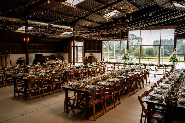 Rustic barn venue featuring elegant wooden tables, string lights, and large windows overlooking lush greenery, perfect for an intimate wedding reception.