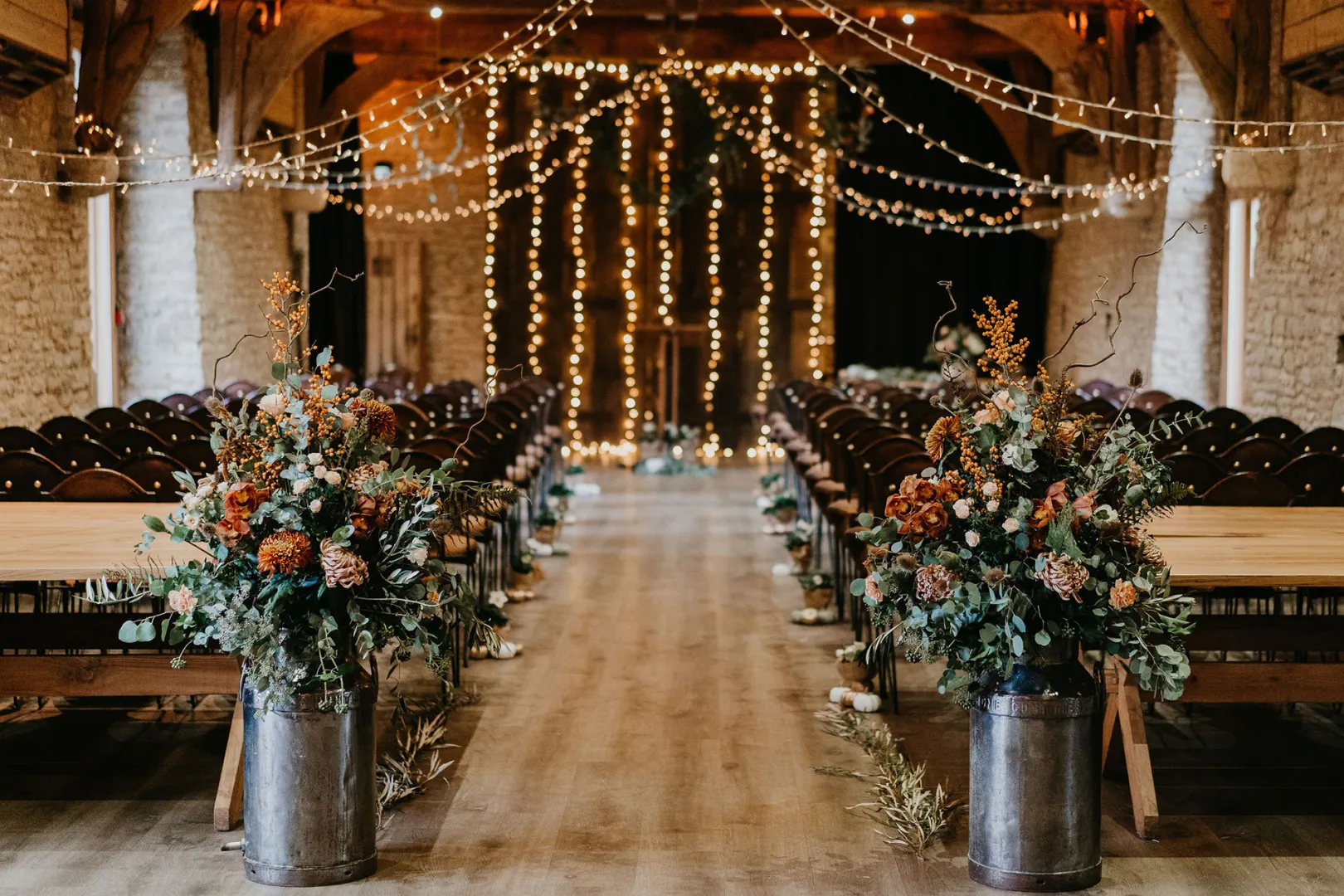 Rustic barn venue with wooden beams, string lights, and floral arrangements lining the aisle for a warm, romantic ceremony atmosphere.