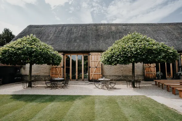 Charming rustic barn with a thatched roof, surrounded by lush greenery and outdoor seating for a picturesque wedding setting.