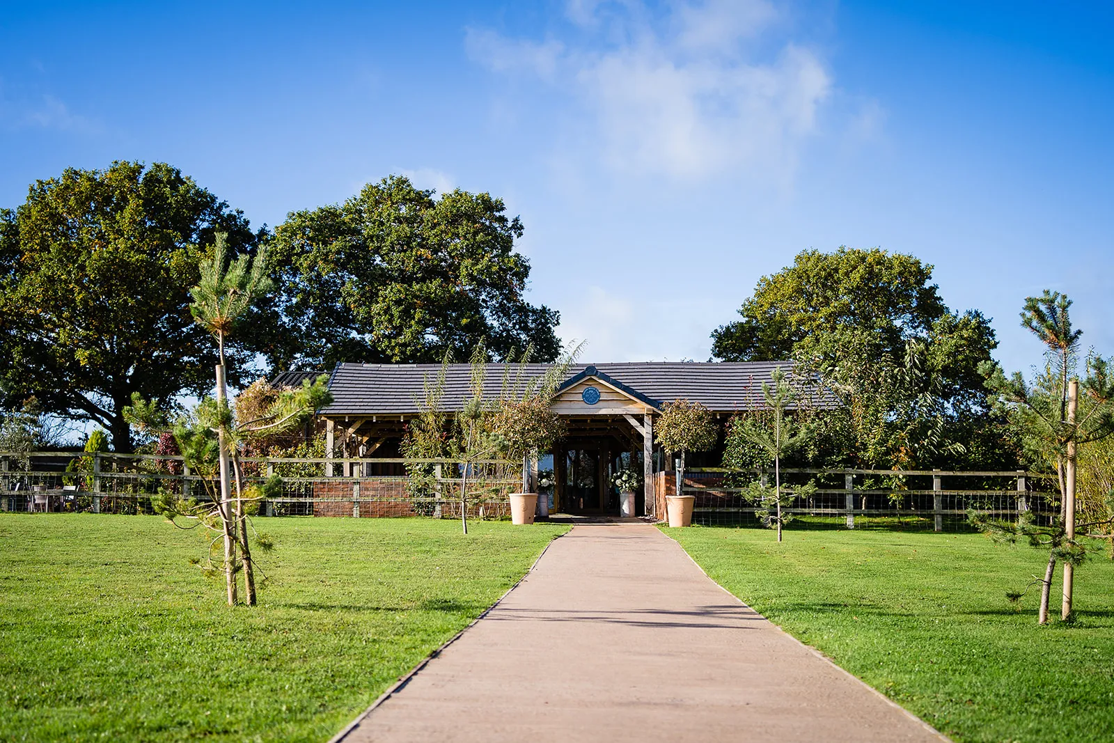 Charming rustic venue entrance with a pathway leading to a cozy building, surrounded by lush greenery and gentle trees. Perfect for outdoor events.